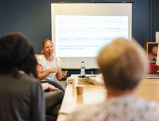 A trainer sits at the front of a classroom speaking in front of a presentation.