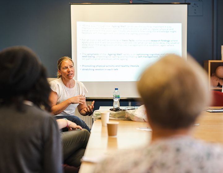 A trainer sits at the front of a classroom speaking in front of a presentation.