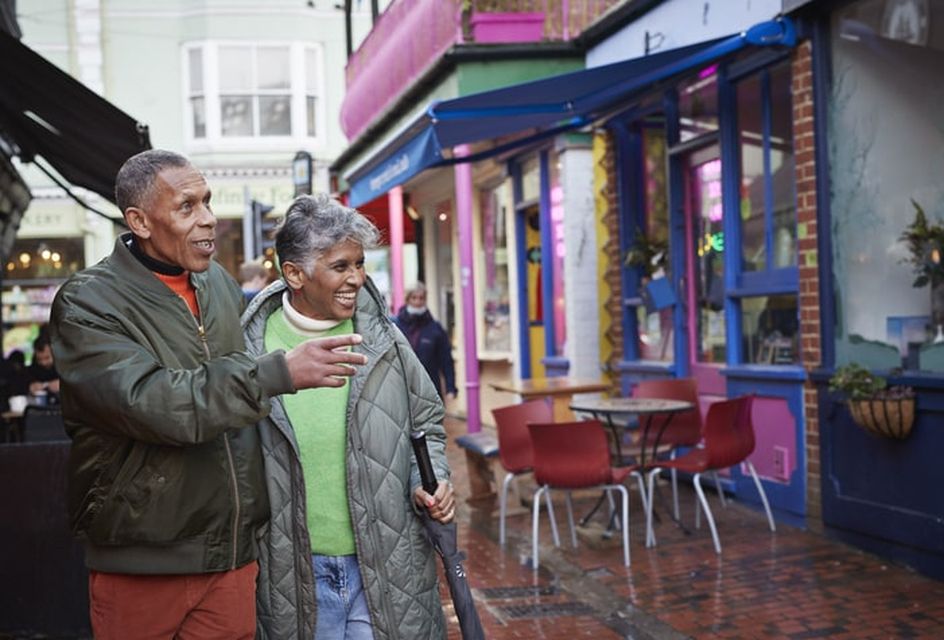 Older man and woman smiling while walking down a British high street on a rainy day with a traditional cafe in shot.