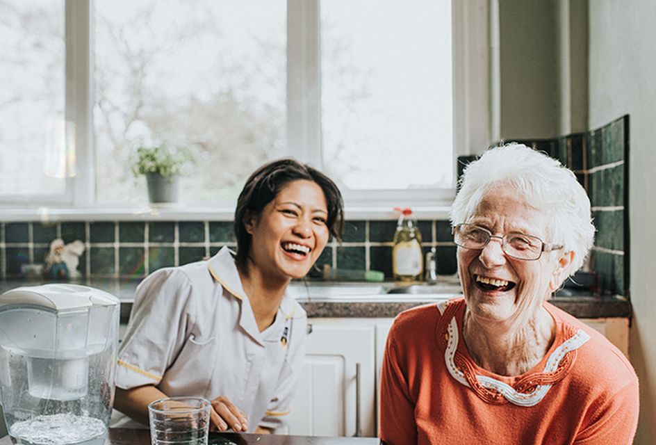 An elderly white woman with grey hair and glasses laughs with her carer, and younger woman with brown hair.