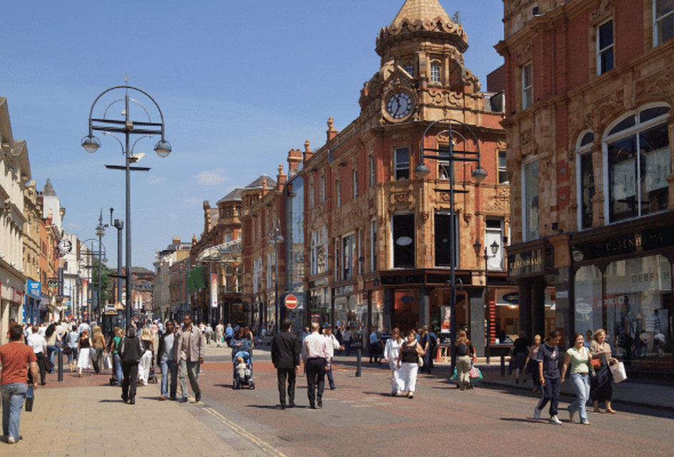A bustling Leeds city centre street with pedestrians, lined with historic buildings featuring ornate architecture and a clock tower