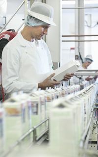 A man in a white coat, hair net and hat inspects bottles of milk in a factory.