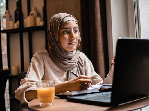 A young muslim woman wearing a hijab studies on a laptop while making notes in a notebook.
