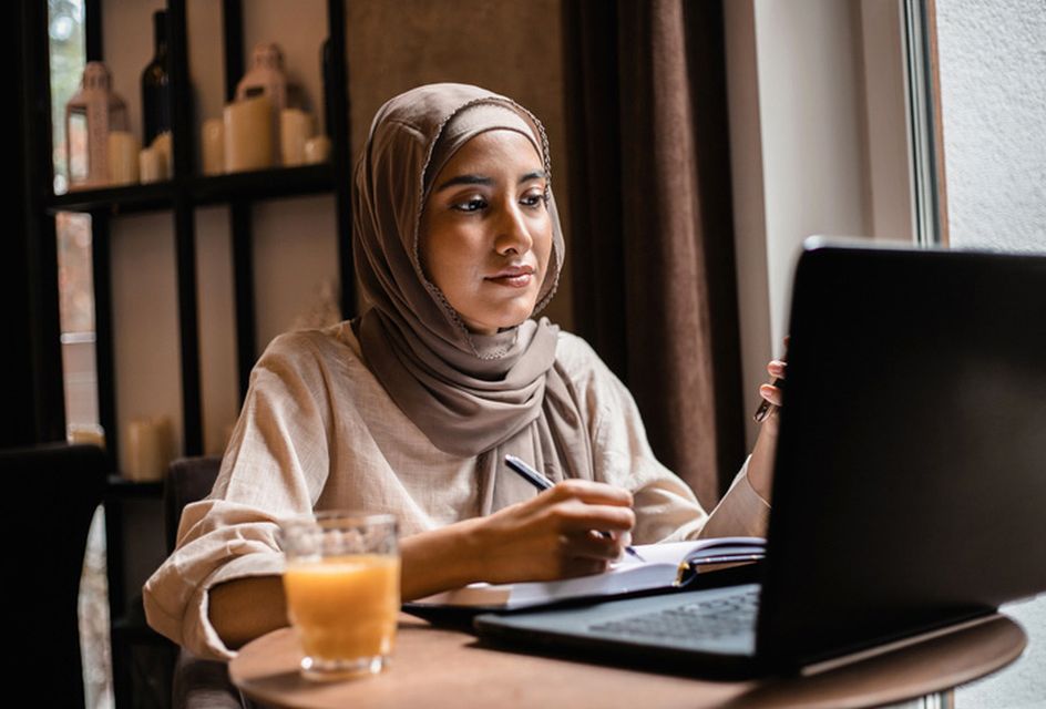 A young muslim woman wearing a hijab studies on a laptop while making notes in a notebook.