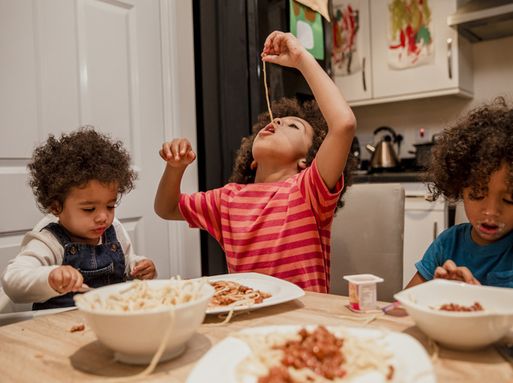 Three young children eating spaghetti at a kitchen table.