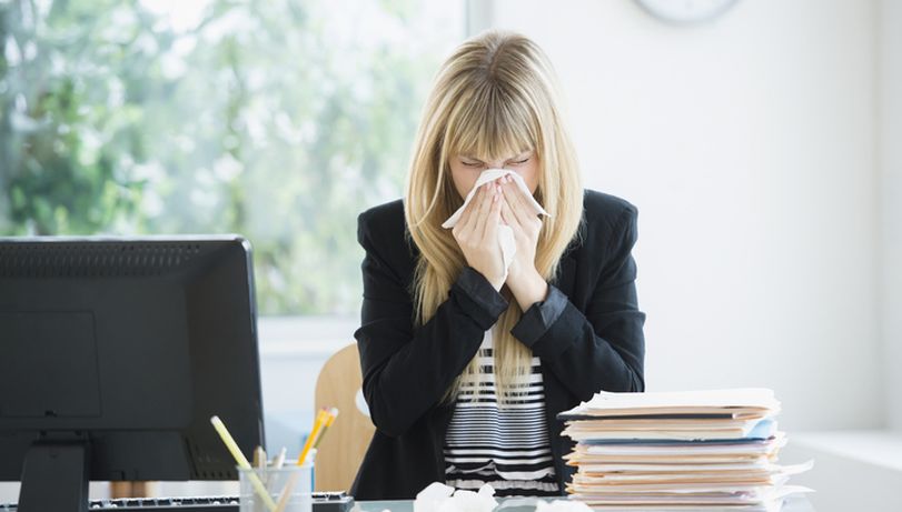 A white woman with blonde hair sneezes into a tissue in an office environment