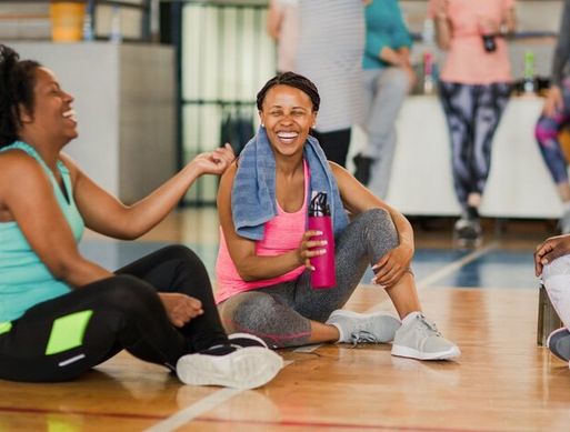Two black women sit on the floor of a gym laughing and talking. They are wearing sports clothes and one has a towel around her neck.