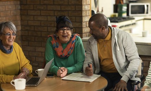 Two women and a man sit at a round table, warmly smiling and engaged in conversation. A laptop and cups are on the table