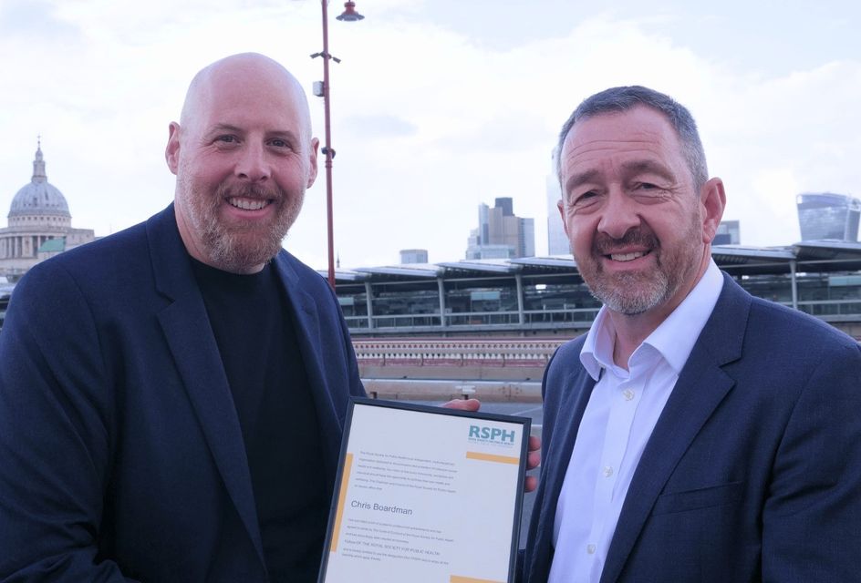 William Roberts and Chris Borardman CBE smiling while holding an RSPH award certificate on Blackfriars bridge, with London landmarks visible in the background