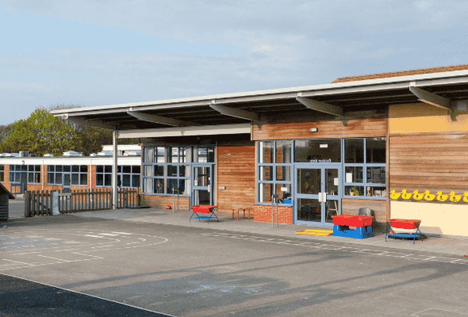 Modern primary school playground with outdoor toys, large windows, and wooden exterior on a sunny day.