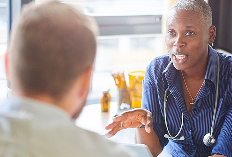 NHS doctor sitting down wearing a stethoscope calmly explaining something to a patient.