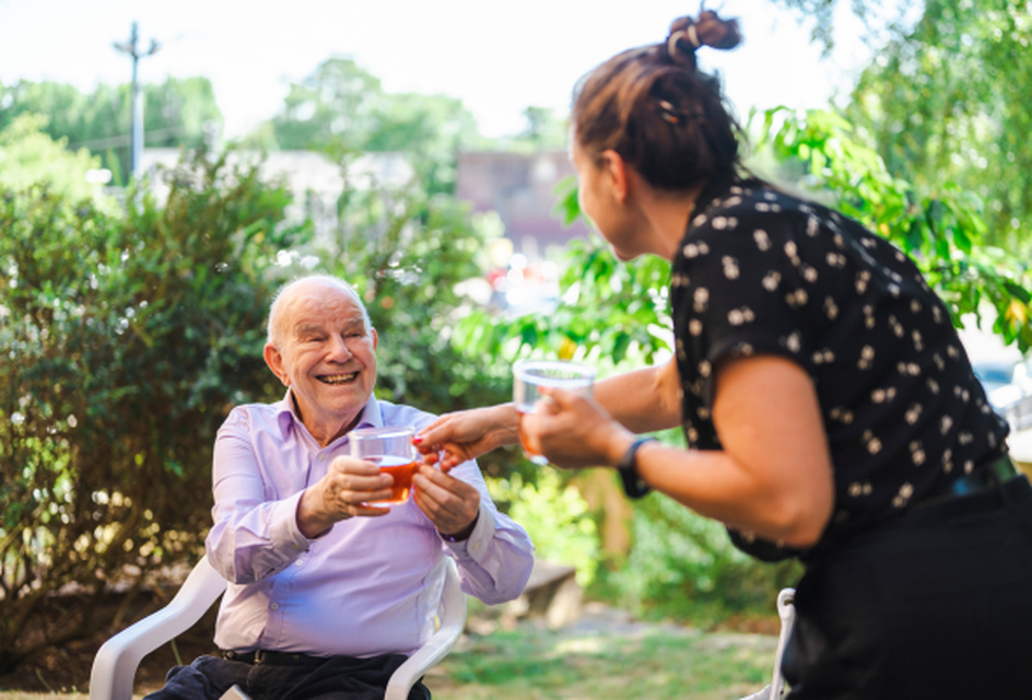 Smiling elderly man receiving a drink from a woman in a sunny garden setting.