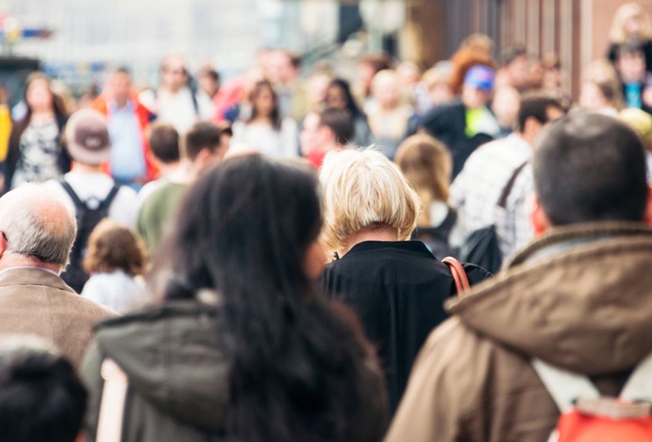 Large crowd of people walking in a busy urban area, viewed from behind.