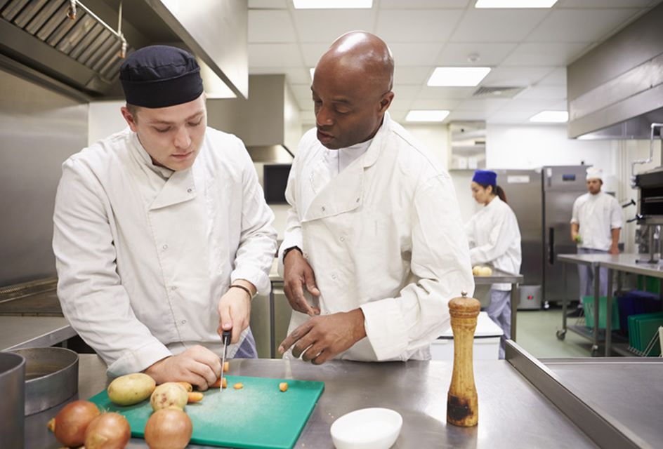 A chef in a white coat supervises another chef as he chops onions.