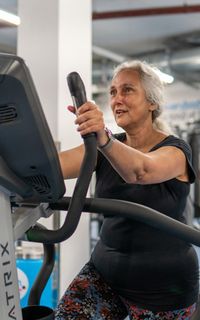 An older woman with grey hair exercises on an elliptical machine in a gym.