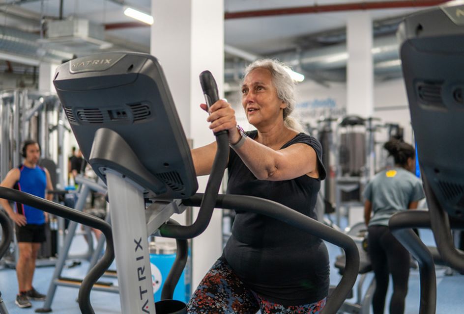 An older woman with grey hair exercises on an elliptical machine in a gym.