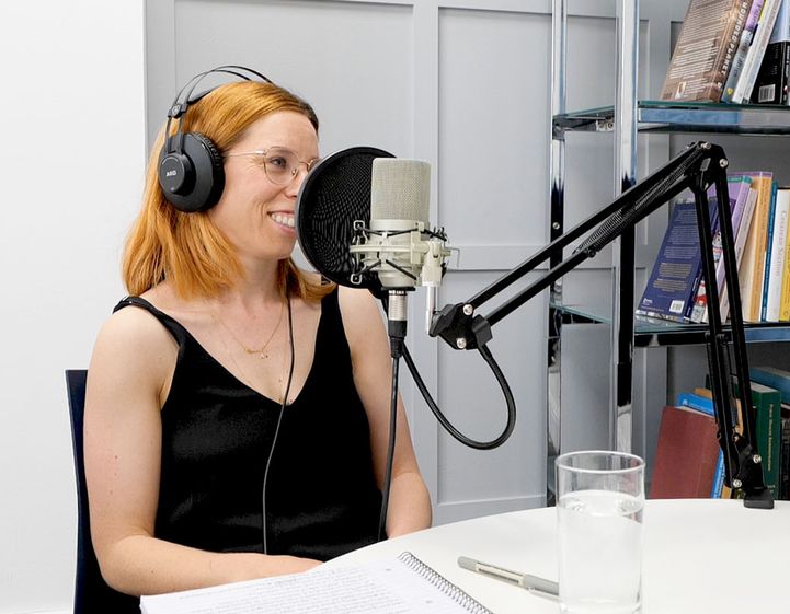 Sara Ogilvie, a white woman with read hair, smiles while recording a podcast. She is wearing headphones and sat behind a microphone.