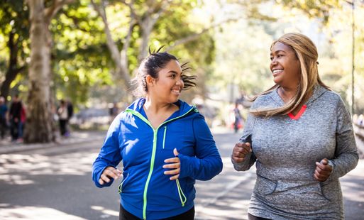 Two women jogging side by side smiling at each other on a sunny day with trees lining the street.