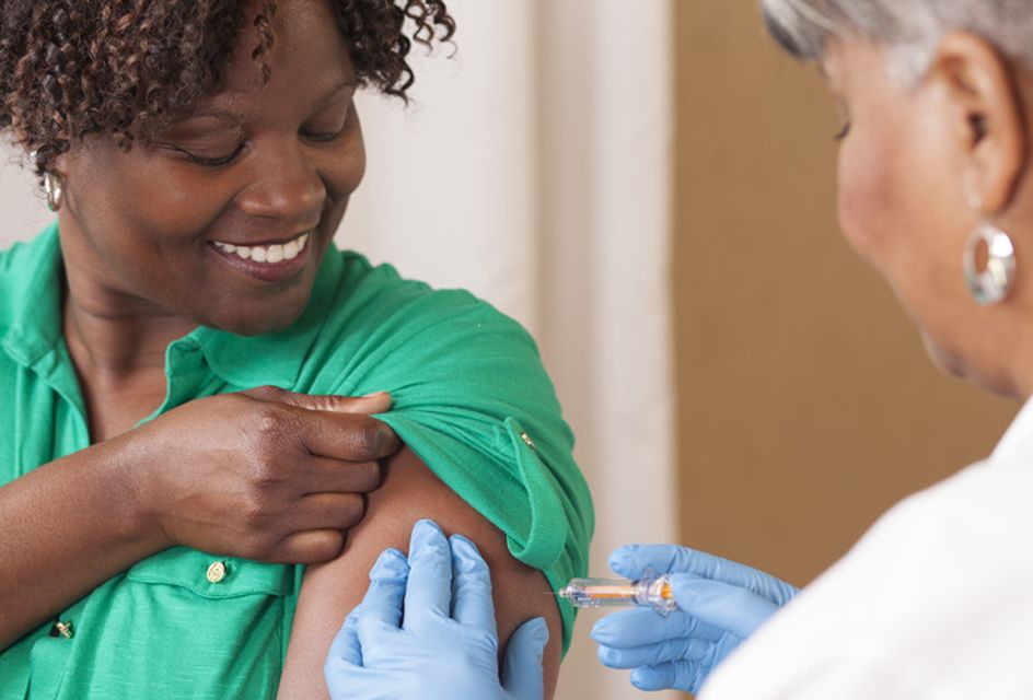 A black woman with short curly hair smiles while getting a vaccine.