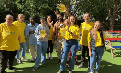 A group of eight North Paddington climate champions stands outdoors on a sunny day, smiling. The background features trees and colourful benches, conveying a cheerful, community-focused atmosphere.
