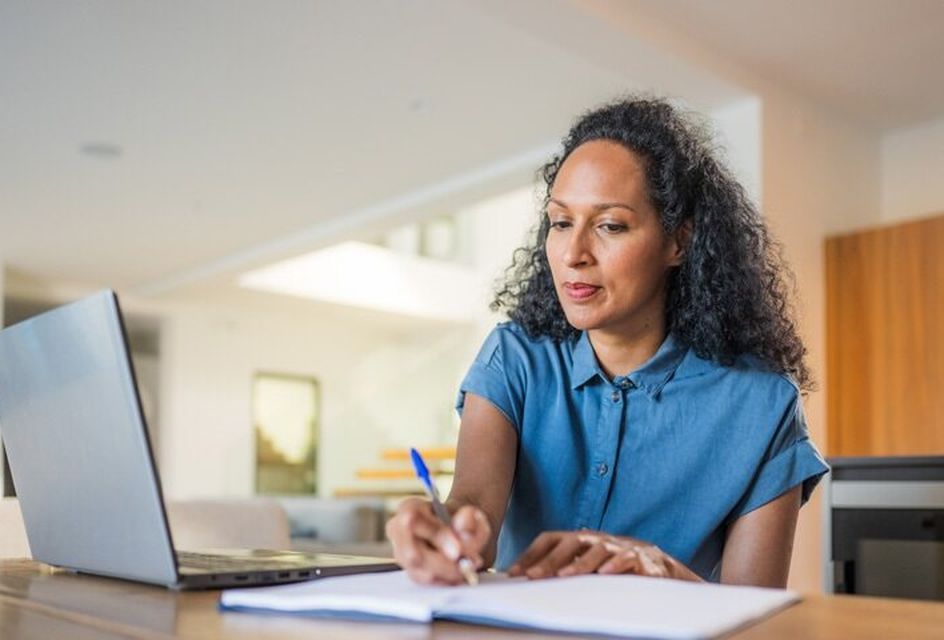 A woman is writing on a notepad with a laptop next to her.