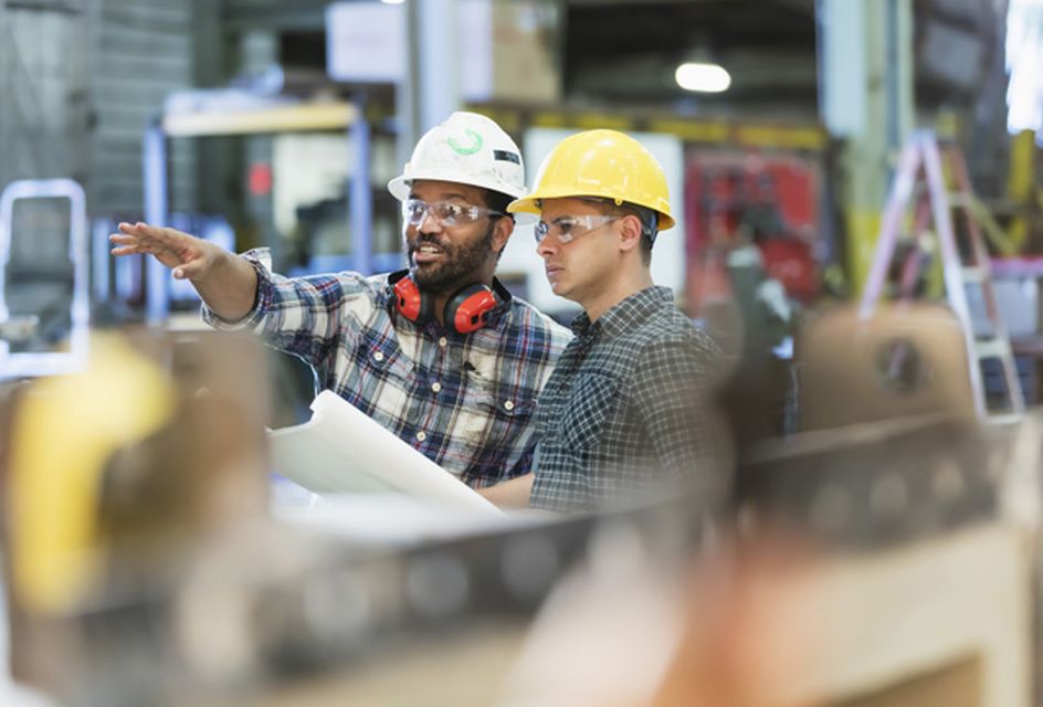 Two man wearing hard hats and safety goggles in a warehouse setting. One of the men is pointing to something out of shot.